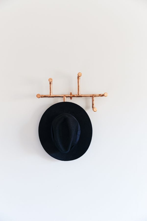 beyaz duvar üzerine monte edilmiş bakır boru aslılık ve askılıkta asılı siyah şapka ön görünüş